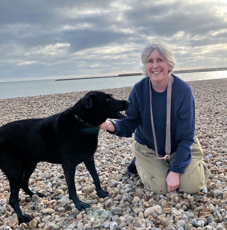 Jane Yates kneeling on the pebbles of Lyme Regis beach beside Oak, her senior black Labrador, with the Cobb and the sea behind them on a grey November afternoon